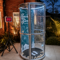 Winter-themed cylindrical glass display labeled 'Snow Store', with falling snowflakes and white balls at the base, next to a brick building; a tripod with a red LED '88' display nearby.