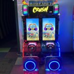 Two-seat arcade racing game cabinet labeled 'Bigfoot Crush' with neon blue wheel rims and bright LED lights.