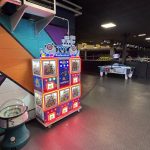 Colorful toy prize vending machine against a teal and purple mural in an indoor arcade.