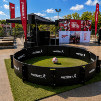 Circular black FASTFEET barrier encloses a small grassy soccer area with a ball in the center at an outdoor event venue and stage behind it.