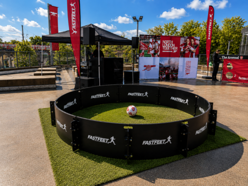 Circular black FASTFEET barrier encloses a small grassy soccer area with a ball in the center at an outdoor event venue and stage behind it.