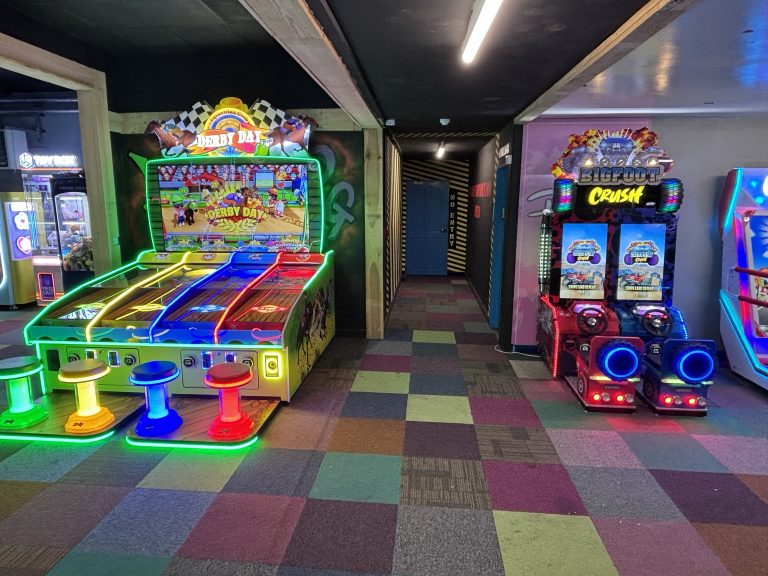 Colorful arcade with neon-lit Derby Day game and stools in the foreground; a Bigfoot Crush machine to the right.